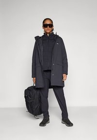 Cappotto lungo nero con cappuccio, indossato sopra una giacca imbottita e pantaloni coordinati. Zaino nero, stivali resistenti e occhiali da sole oversize.