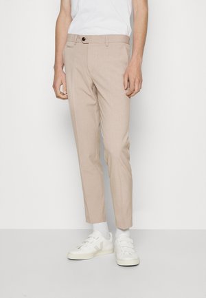 Mann trägt beige maßgeschneiderte Hose, weiße Socken, weiße Klettverschluss-Sneakers und ein weißes T-Shirt vor einem schlichten hellen Hintergrund.