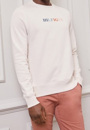 Man die een witte lange mouw sweatshirt draagt met het "HILFIGER"-logo en zalmroze broek, staand tegen een witte gegroefde muur.