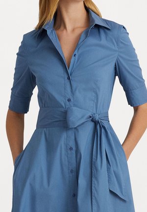 Femme portant une robe chemise bleue à manches courtes avec une ceinture nouée à la taille, mains dans les poches, sur un fond uni.