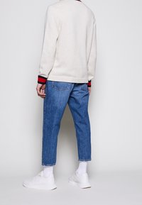 Pull crème avec des poignets rayés rouges et noirs, associé à un jean bleu denim ample et des baskets blanches, vu de l'arrière.