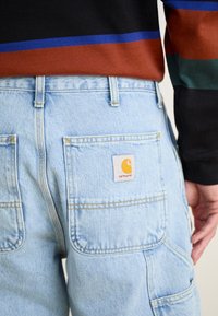 Pantalones de mezclilla azul claro con dos bolsillos traseros, costuras amarillas y una etiqueta color beige con un logo. Llevados con una blusa de rayas multicolores.
