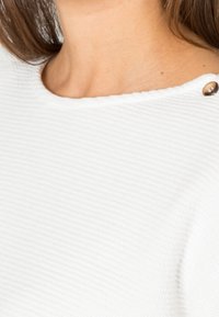 White ribbed sweater featuring a rounded neckline, textured fabric, and a decorative button accent near the shoulder.