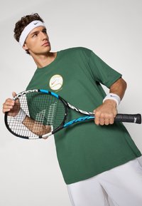 Grøn bomulds-T-shirt med rundt Nike-logo; hvid svedbånd på hovedet og håndleddet; sort og blå tennisracket med en tekstureret håndtag.