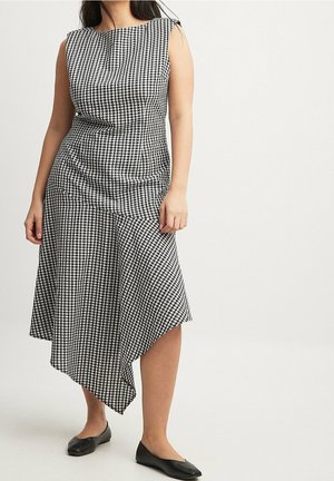 Sort-hvid gingham midi-kjole med en tætsiddende overdel, draperede detaljer, asymmetrisk kant og ærmeløs design. Sorte flade sko fuldender looket.