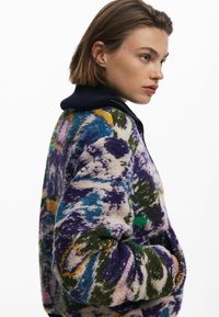 Giacca in pile colorata con un motivo vorticoso in viola, verde e blu, con colletto nero e chiusura con zip. Tessuto morbido e strutturato.