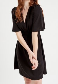 Robe noire avec manches courtes volantées, encolure en V et taille élastique. Tissu doux avec une coupe décontractée s'étendant jusqu'au milieu des cuisses.