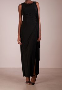 Svart maxiklänning med draperad design, ärmlös silhuett, asymmetrisk skärning och hög sidoklyfta. Slät, stretchig tyg. Svarta klackar ingår.