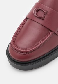 Mocassin en cuir bordeaux avec un accent de logo brillant, des détails cousus et une semelle en caoutchouc texturé noire. Design à bout rond et plat.