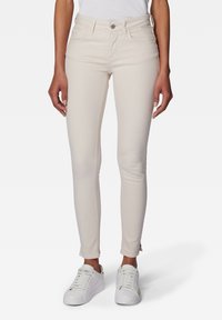 Jean skinny beige clair avec une taille mi-haute, des poches avant et un ourlet légèrement effiloché ; porté avec des baskets blanches.