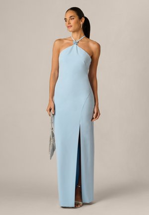 Kobieta w jasnoniebieskiej sukni na szyję z rozcięciem na udzie trzyma srebrną torebkę z siateczki, ma na sobie srebrne szpilki i kolczyki na neutralnym tle.