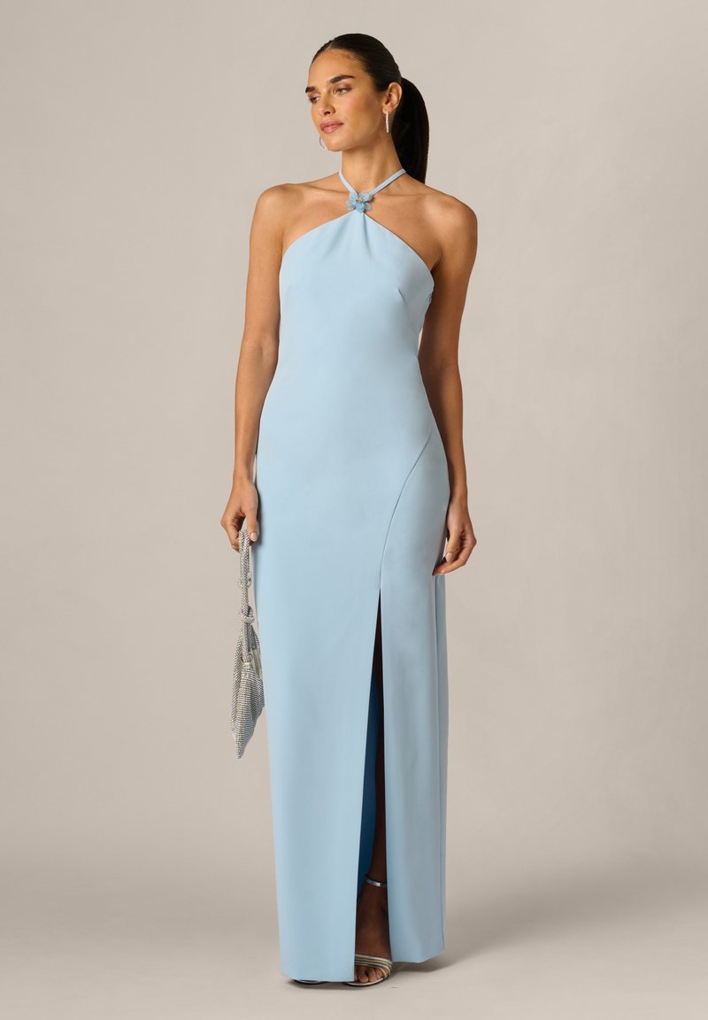 Žena v světle modrých šatech s halter výstřihem a rozparkem na stehně drží stříbrnou síťovanou kabelku, navíc má na sobě stříbrné podpatky a náušnice na neutrálním pozadí.