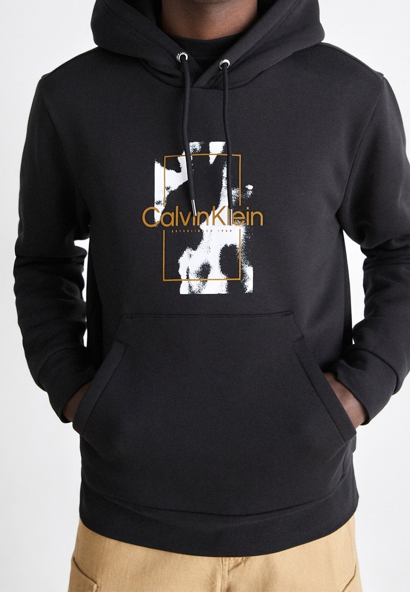 Felpa nera con cappuccio e tasca frontale, coulisse regolabile e stampa grafica con "Calvin Klein" e un motivo bianco e oro.