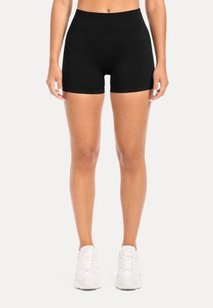 Femme portant un short noir moulant et des baskets blanches, debout, les bras détendus le long du corps sur un fond uni.