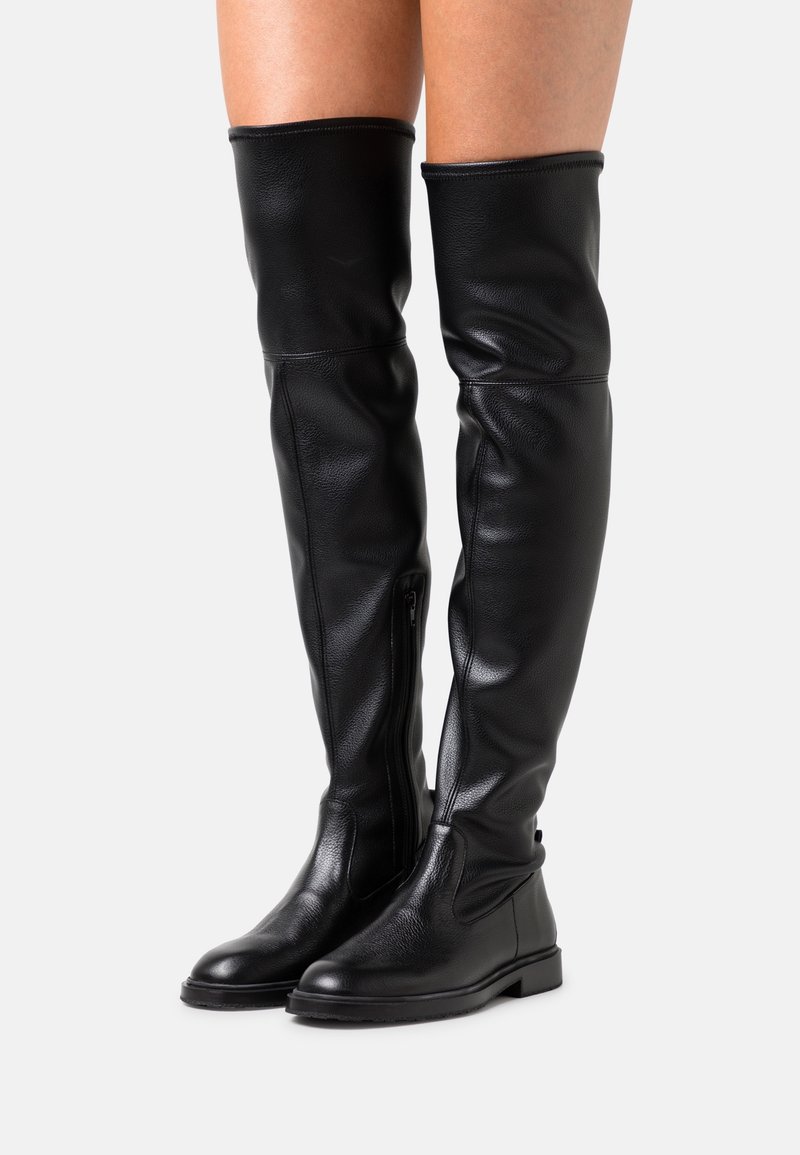 Bottes plates en cuir noir montant au-dessus du genou avec fermetures éclair latérales portées sur des jambes nues, sur fond blanc.