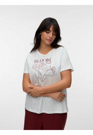 Kvinde med mørkt hår iført en hvid T-shirt med blomstertegning og teksten "BLOOM", stående med armene over kors på en ensfarvet lys baggrund.