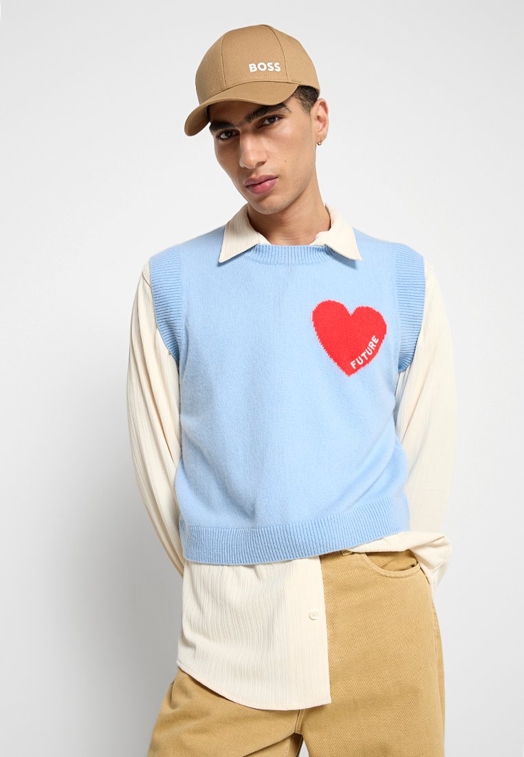 Gilet en tricot bleu clair avec un graphique de cœur rouge et le texte "FUTURE", superposé sur une chemise à col crème, associé à un pantalon beige et une casquette.
