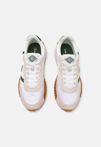 Sneakers met een witte synthetische bovenkant, accenten van beige suède, groene strepen en een getextureerde oranje rubberen zool. Lacoste-logo aan de zijkant.
