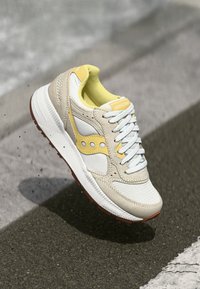 Light beige sneaker with yellow accents and white mesh. Features a textured design, white laces, and a rubber sole with brown traction.