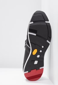 Semelle de basket noire avec talon rouge, accents blancs, motif géométrique de la semelle et logo Vibram jaune près de la voûte plantaire.