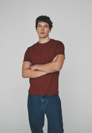 Ung mand med mørkt krøllet hår står med armene krydset iført en vinrød t-shirt og blå jeans foran en ensartet lys baggrund.