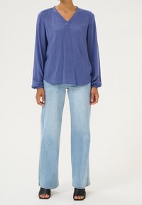 Blouse bleu à manches longues avec un décolleté en V, présentant un détail froncé à l'épaule et une coupe décontractée, associée à un jean à jambes larges bleu clair et des talons noirs à bout ouvert.