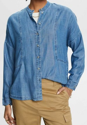 Personne portant une chemise en jean bleu clair à boutons avec un col mandarin, un pantalon cargo beige et des bijoux en or à la main et au poignet.