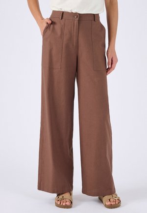 Mujer vistiendo pantalones marrones de pierna ancha con bolsillos aplicados, una camiseta blanca y sandalias beige con hebilla, de pie con una mano en el bolsillo.