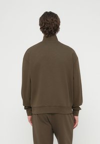 Καφέ φούτερ με ψηλό γιακά, φαρδιά εφαρμογή, ριμπ μανσέτες και τελείωμα. Μαλακή υφή, μινιμαλιστικός σχεδιασμός χωρίς ορατά σχέδια ή διακοσμητικά στοιχεία.