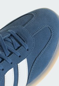 Plave suède tenisice s bijelim kožnim prugama, ravnim vezicama i teksturiranom površinom. Gumeni potplat i detalji šivanja su vidljivi.