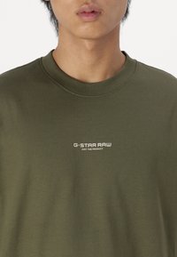 Olive zelená bavlněná mikina s kulatým výstřihem a krátkými rukávy. Na hrudi se nachází bílý text s logem. Hladká textura.