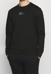 Sudadera negra de tejido suave, con mangas largas, cuello redondo y un logo blanco en el pecho. Puños y dobladillo acanalados.
