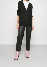 Musta neuletakki, jossa on suuret napit, V-muotoinen kaula-aukko, ribattu hihansuu ja taskut, yhdistettynä mustiin nahkahousuihin ja vaaleanpunaisiin korokepolkimiin.