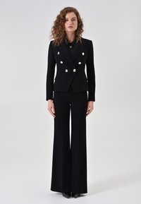 Costume deux pièces noir comprenant un blazer croisé à boutons métalliques, des revers nets, et un pantalon à jambe large avec une texture en chevrons.