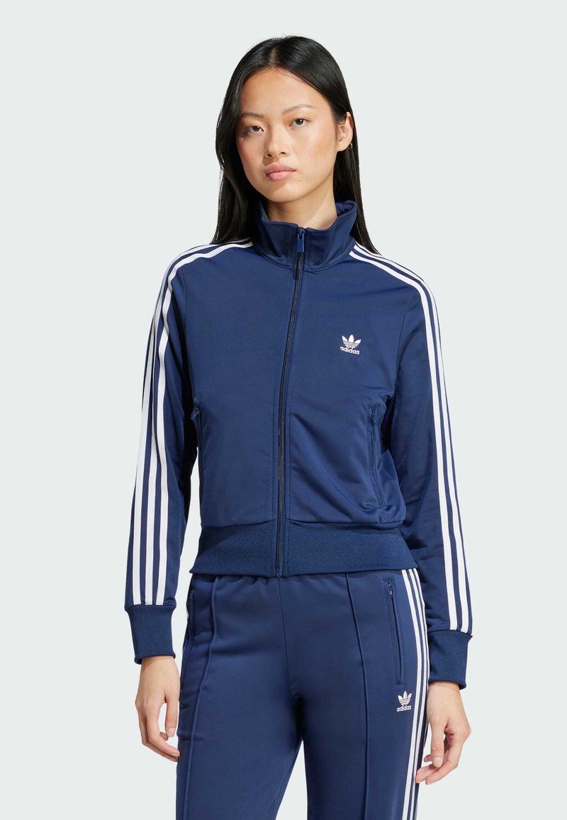 Dunkelblaues Trainingsjacke mit weißen drei Streifen-Akzenten, hohem Kragen, Reißverschluss und einem kleinen Adidas-Logo auf der Brust. Weicher Stoff.