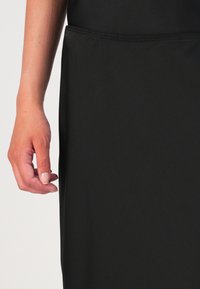 Musta mekko, joka on valmistettu sileästä kankaasta, siinä on istuva vyötärö ja osittain näkyvä käsi lepäämässä sivulla. Saumayksityiskohtia on läsnä.