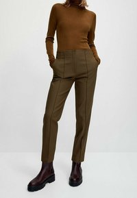 Brown ribbed turtleneck; olive green high-waisted trousers with vertical seams; dark burgundy ankle boots with textured soles.