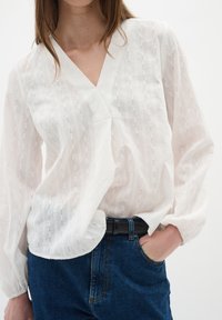 Blusa bianca a maniche lunghe con scollatura a V e strisce testurizzate. Abbinata a jeans in denim blu e una cintura nera, presenta una vestibilità comoda.
