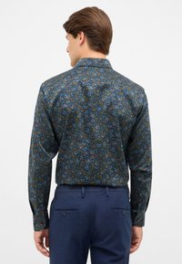 Donker gestippeld shirt met een bloemenpatroon in blauwe, oranje en groene tinten; lange mouwen en een opgevouwen kraag. Gepaard met marineblauwe broeken.