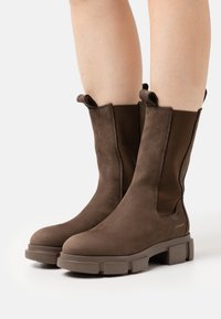 Bottes chelsea brunes en cuir texturé avec panneaux élastiques latéraux, bout rond et semelle en caoutchouc épaisse. Comprend des languettes pour un enfilage facile.