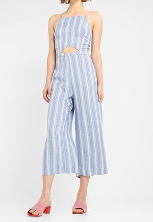 Combinaison rayée bleu et blanc avec une découpe à la taille. Dotée de fines bretelles et d'un pantalon large raccourci. Associée à des sandales à talons blocs roses.