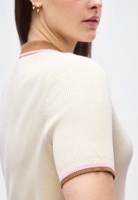 Ženska, oblečena v kremno kratko majico z rebrastim vzorcem, rjavim in roza okrasom na ovratniku in rokavu, prikazana od ramen do brade.