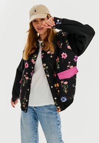 Czarna kurtka w kwiatowy wzór z nadmuchanymi rękawami, różowymi akcentami na kieszeniach i srebrnymi zatrzaskami; noszona na białej koszuli i niebieskich dżinsach.