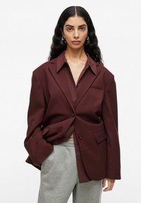 Kvinna med långt mörkt hår, bär stora hoops örhängen, en överdimensionerad mörk burgunderblazer, matchande skjorta och ljusgrå sweatpants, vit bakgrund.