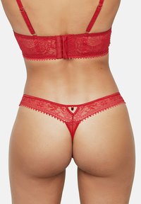 Ensemble de lingerie en dentelle rouge comprenant un soutien-gorge avec bretelles ajustables et fermeture à agrafes, ainsi qu'un string avec un petit détail rouge.