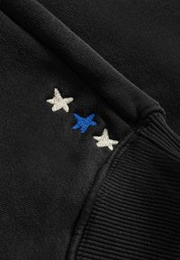 Czarna tkanina o prążkowanej teksturze, z trzema haftowanymi gwiazdami: jedną niebieską i dwiema białymi, ustawionymi w poziomym rzędzie.