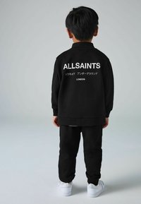 Melnas zip-up sporta jaka ar "ALLSAINTS" tekstu aizmugurē, saskaņota ar melniem biksēm un baltām sporta apaviem. Gluda auduma, brīva piegriezuma.