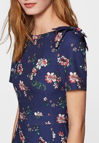 Laivansinis kukkamekko lyhyillä hihoilla, pehmeästä kankaasta, jossa valkoisia, vaaleanpunaisia ja vihreitä kukkakuoseja, sekä nauhakoristeet olkapäillä.