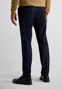 Man in een donkerblauw geruit broek, bruine instappers en een camelgekleurde trui, staand tegen een effen lichte achtergrond.