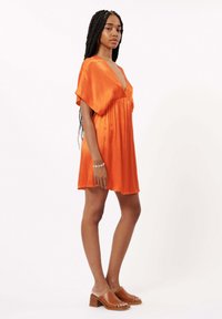Kurzes oranges Satinkleid mit tiefem V-Ausschnitt und kurzen Ärmeln, mit einem lockeren Schnitt. Kombiniert mit braunen Absatzmules.